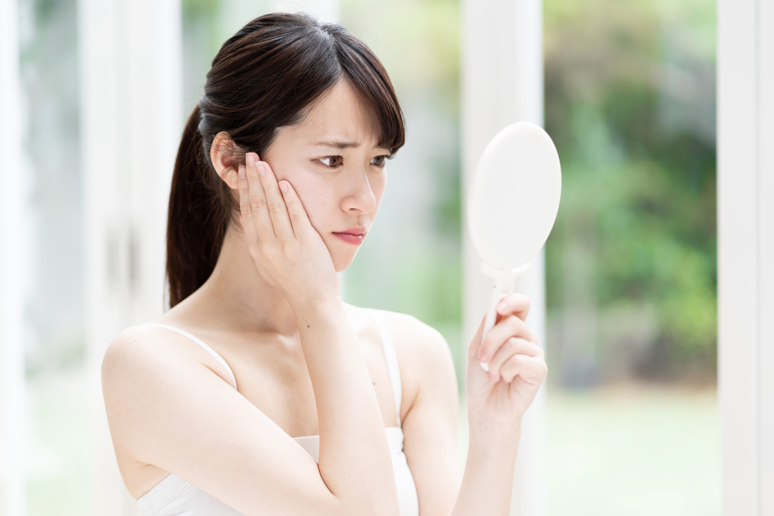 フェイスラインのたるみや肌の悩みを、鏡を見ながら心配そうに確認する女性